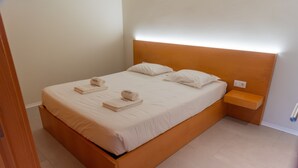 Ein ordentlich bezogenes Bett mit Holz-Kopfstück, einem Nachttisch und einer Wandsteckdose.