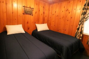 A room with two beds, wooden walls, a framed picture, and a window with curtains.