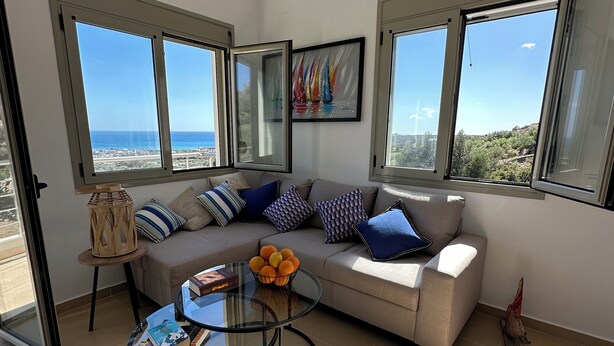 A modern living room with a beige sofa, a glass coffee table, and a view of the sea through large windows.