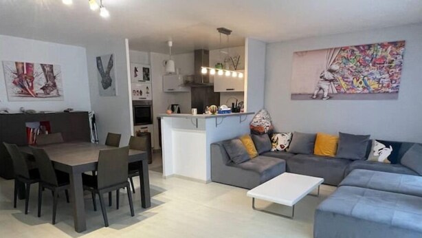 A modern living room with a grey sofa, a dining table with chairs, and a kitchen area in the background. - Appartement F4 Proche du Stade de France (Saint-Denis)