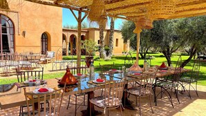 An outdoor dining area with tables and chairs, a thatched roof, and a view of a building with arches.