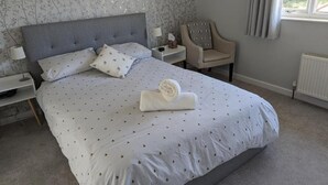 A neatly made bed with a grey headboard, a bedside table with a lamp, a chair, and a window with curtains. - Little Barham B&B (Braintree)