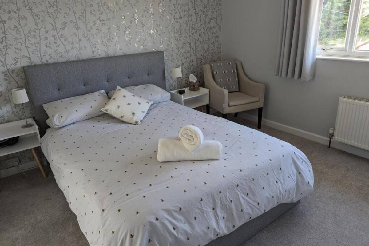 A neatly made bed with a grey headboard, a bedside table with a lamp, a chair, and a window with curtains.