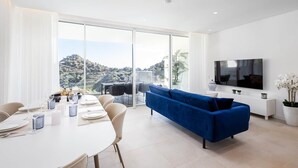 A modern living room with a blue sofa, a dining area with a white table, and a large TV on the wall. - RDR304 - Stunning 2 bedroom Palo Alto, pool, gym + more! (Marbella)