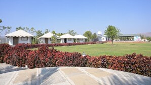 A row of white tents with a flat roof, surrounded by a manicured garden with red foliage, set against a clear blue sky. - Stone Water Resort  (Nashik)