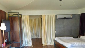 A hotel room with a bed, a ceiling fan, curtains, a desk with a lamp, and an air conditioning unit. - Stone Water Resort  (Nashik)