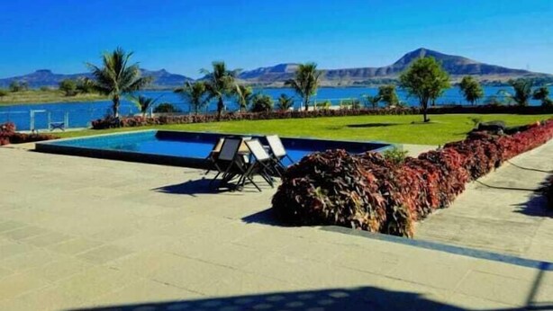 A swimming pool with lounge chairs, a landscaped area with palm trees, and a view of mountains and a body of water. - Stone Water Resort  (Nashik)