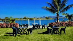 An outdoor seating area with tables and chairs, a grassy lawn, and a view of a lake with mountains in the distance. - Stone Water Resort  (Nashik)
