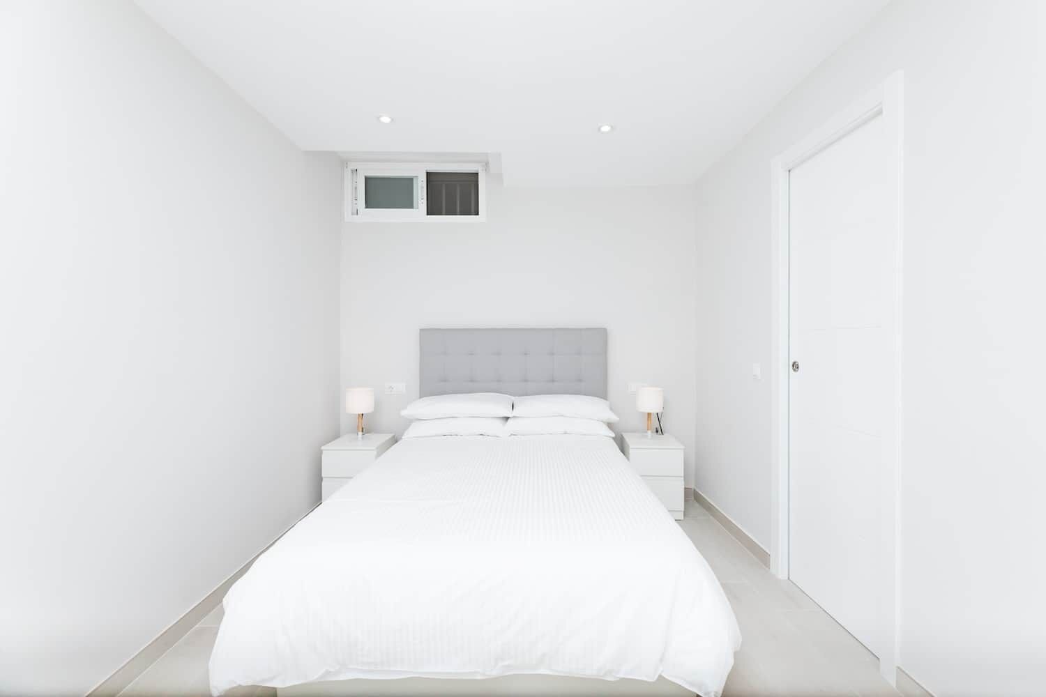 Ein schlicht eingerichtetes Schlafzimmer mit einem großen Bett, zwei Nachttischen mit Lampen, einem Fenster und einer Tür.