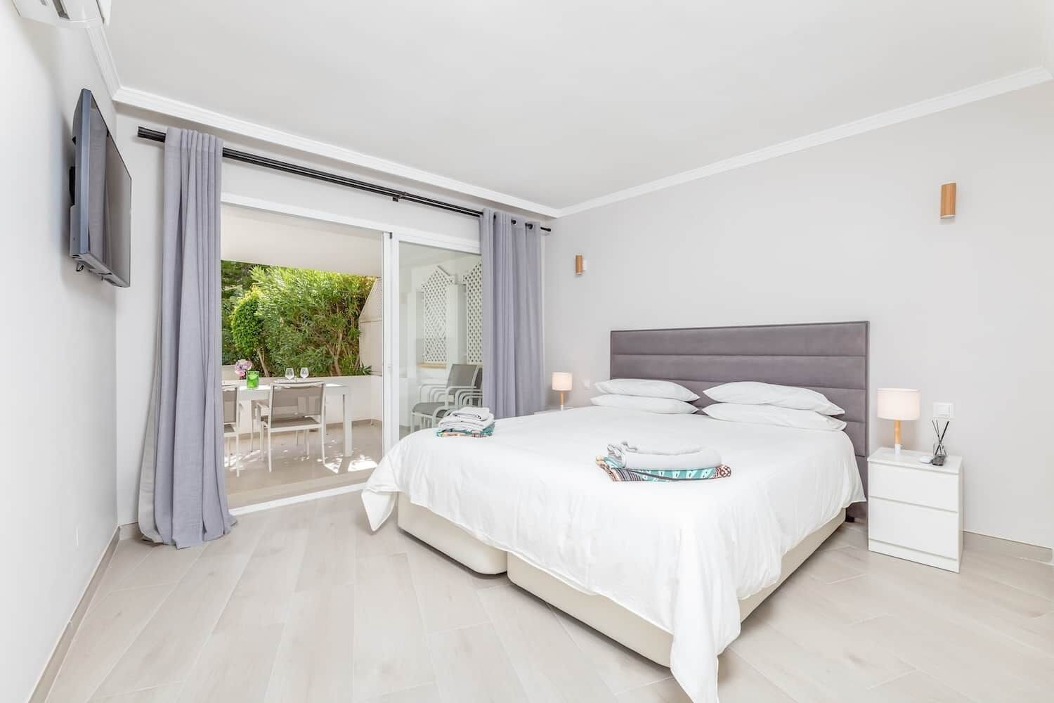 Une chambre moderne avec un grand lit, une table de chevet et une vue sur un balcon aménagé avec des sièges d’extérieur.