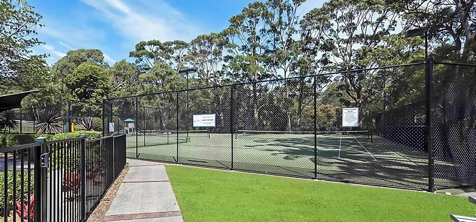 Una cancha de tenis cercada, con un camino que la lleva a ella, rodeada de árboles y cielo despejado.
