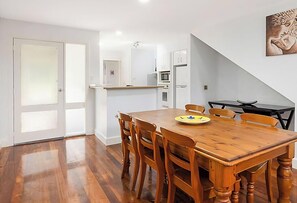 A dining area with a wooden table and chairs, a small kitchen in the background, and a door leading outside.