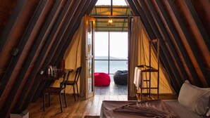A cozy, triangular room with a bed, a red bean bag, a wooden table with chairs, and a view of the outdoors through a large window. - The Backyard Blangoda (Balangoda)