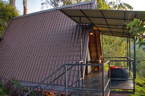 A rustic cabin with a metal roof, a wooden deck, and a staircase leading to a covered porch. - The Backyard Blangoda (Balangoda)