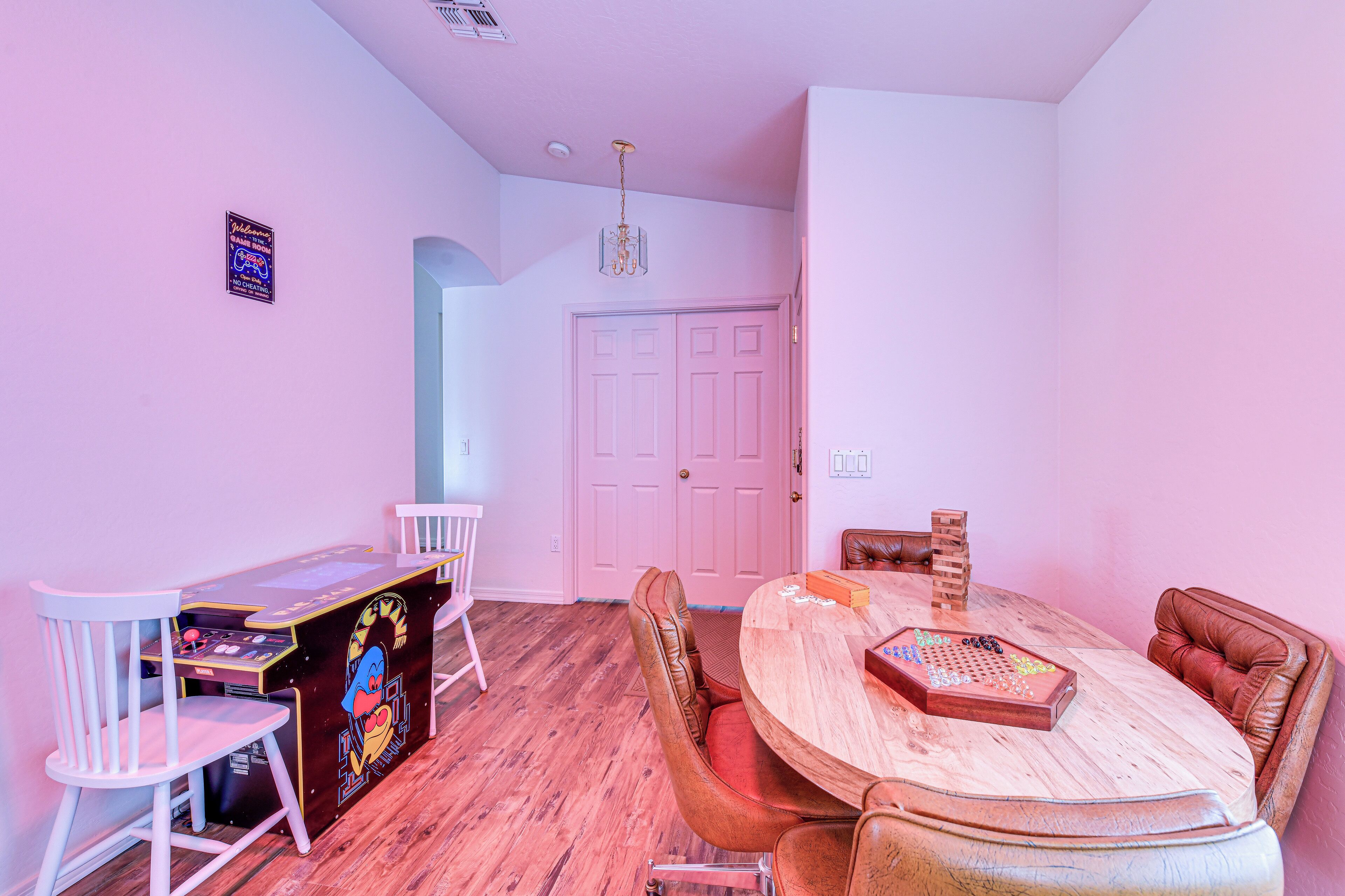 Ein Zimmer mit einem Holzesstisch, Stühlen, einer kleinen Spielecke mit einem Spielautomaten und einer pinken Tür.