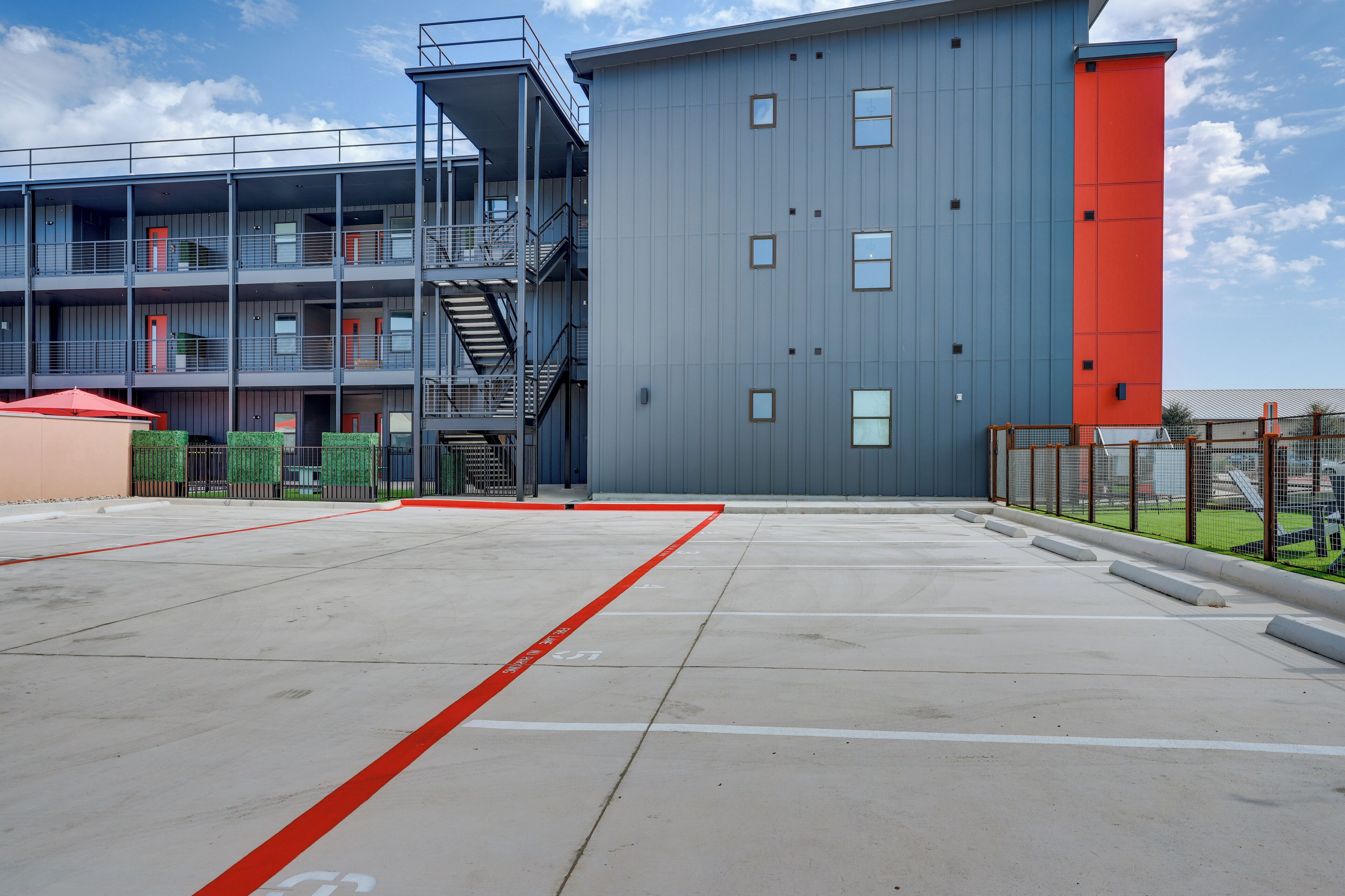 Un immeuble à plusieurs étages, doté d’une façade rouge et grise, d’un espace de stationnement avec des places délimitées et d’un escalier menant à un niveau supérieur.
