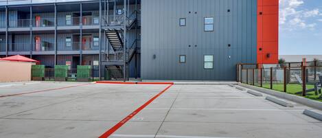 Un edificio a più piani con facciata rossa e grigia, un'area di parcheggio con posti delimitati e una scala che conduce a un livello superiore.