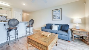 A living room with a blue sofa, two black chairs, a wooden coffee table with a chessboard, a ceiling fan, and a lamp on a side table. - Walk to Beach: North Myrtle Beach Condo w/ Sunroom (North Myrtle Beach)