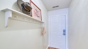 A hallway with a white door, a wooden floor, and a wall-mounted shelf with hats and a framed picture. - Walk to Beach: North Myrtle Beach Condo w/ Sunroom (North Myrtle Beach)
