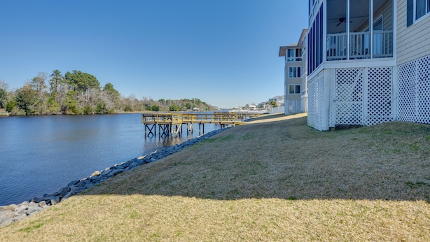 A waterfront property with a wooden dock extending into a lake, a grassy area, and a building with a balcony and lattice design. - Walk to Beach: North Myrtle Beach Condo w/ Sunroom (North Myrtle Beach)