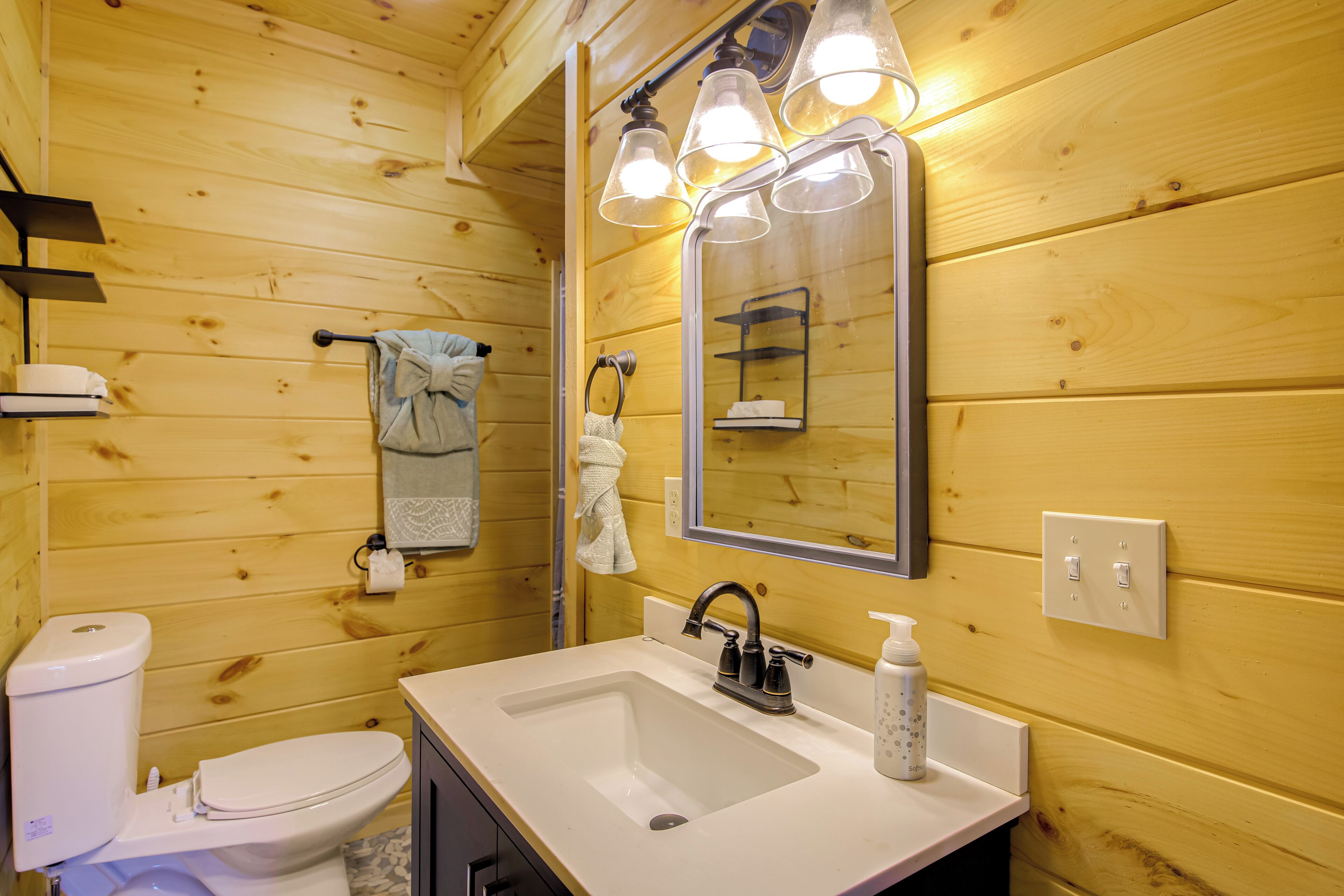 A wooden cabin bathroom with a white sink, a mirror with lights, a toilet, and a towel rack.