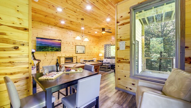 A wooden cabin interior with a dining area, a sofa, and a large window.