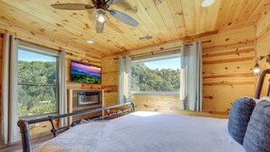 A bedroom with a large bed, a ceiling fan, a TV mounted on the wall, and a view of the forest through the windows.