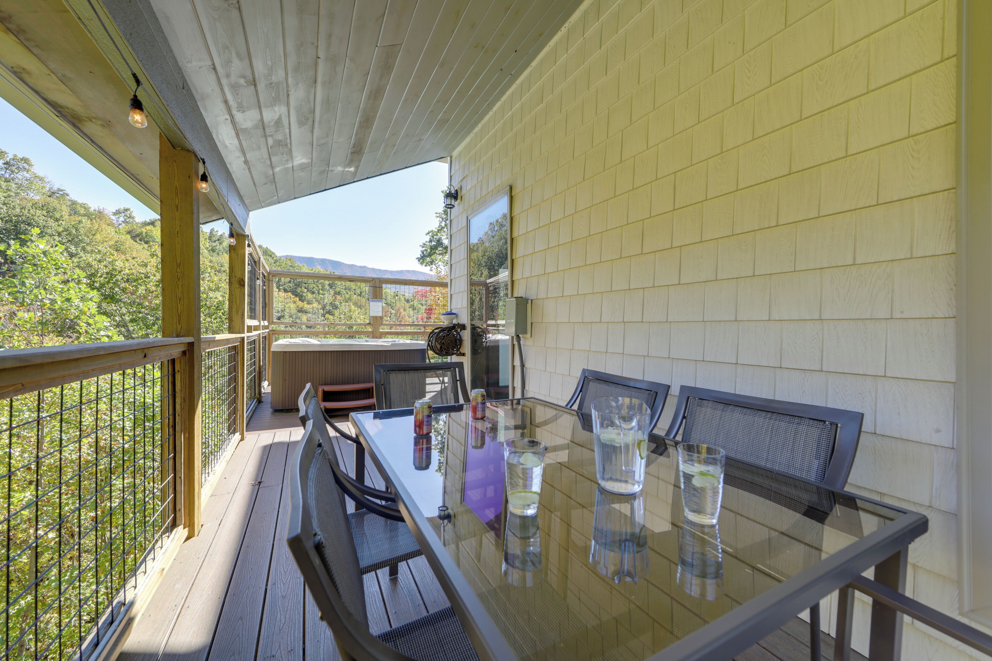 A glass-top table with four chairs, a wooden deck, and a view of trees and mountains.