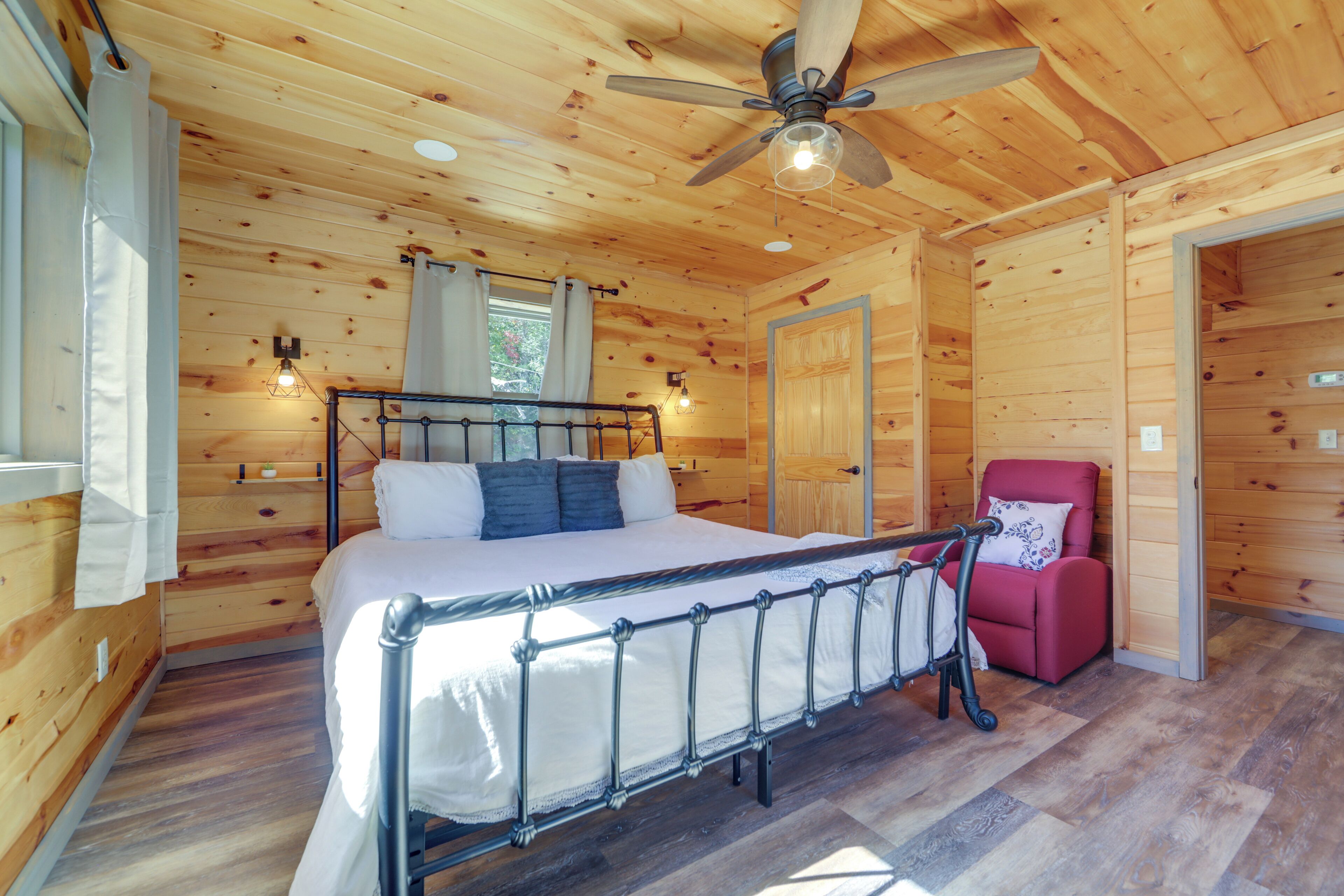 A wooden cabin bedroom with a bed, a red chair, a ceiling fan, and a window with curtains.