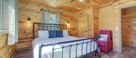 A wooden cabin bedroom with a bed, a red chair, a ceiling fan, and a window with curtains.