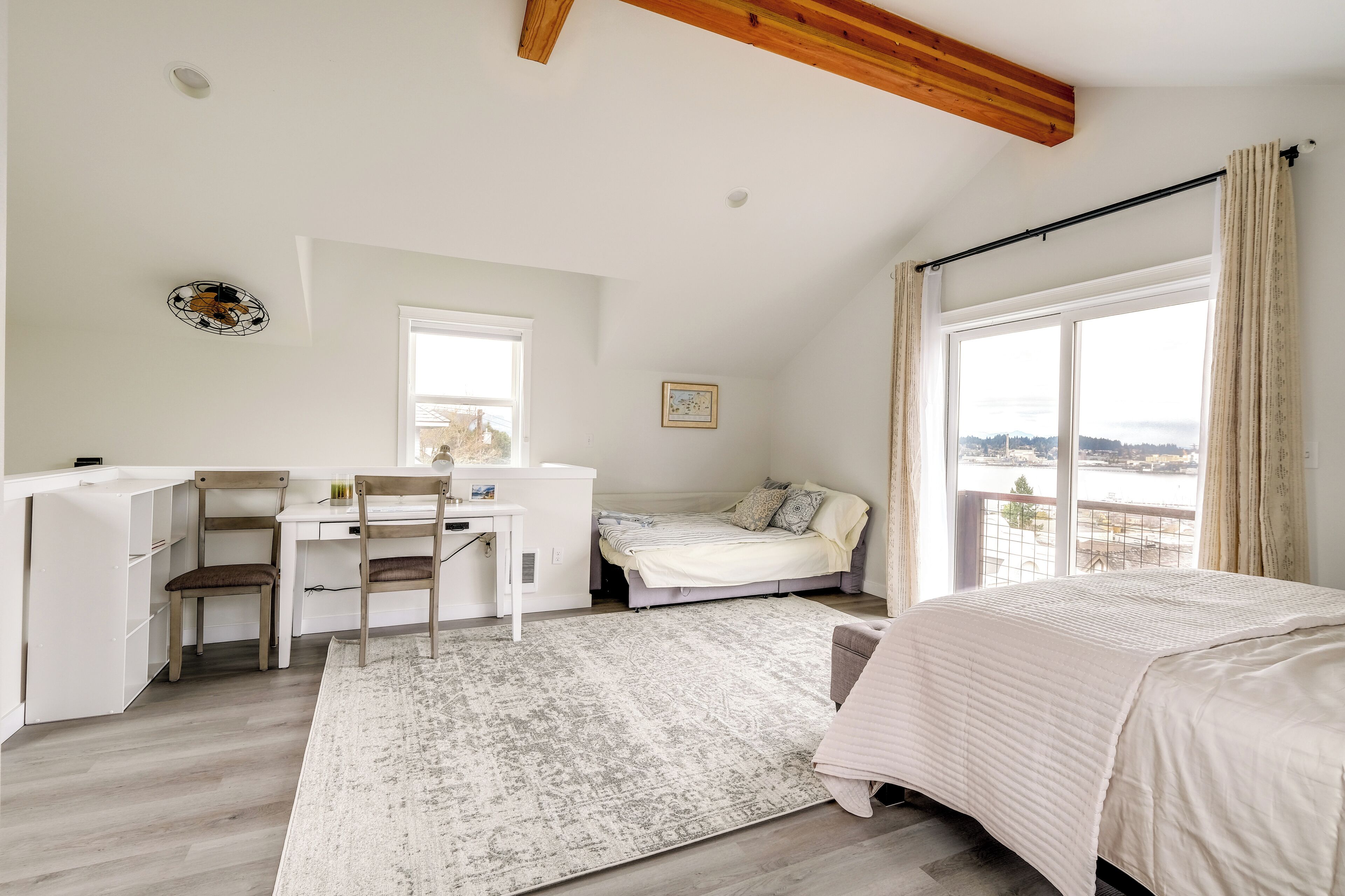 A bedroom with a bed, a desk, and a chair, a large window with curtains, and a view of the outdoors.