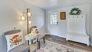 A room with a grey armchair, a small round table, a patterned rug, a white bench, a wall-mounted light fixture, and a door with a wreath. - New Blaine Getaway - Walk to Lake Dardanelle! (New Blaine)