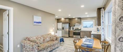 A living room with a sofa, a dining table with chairs, a kitchen area with appliances, and a view of the outdoors.