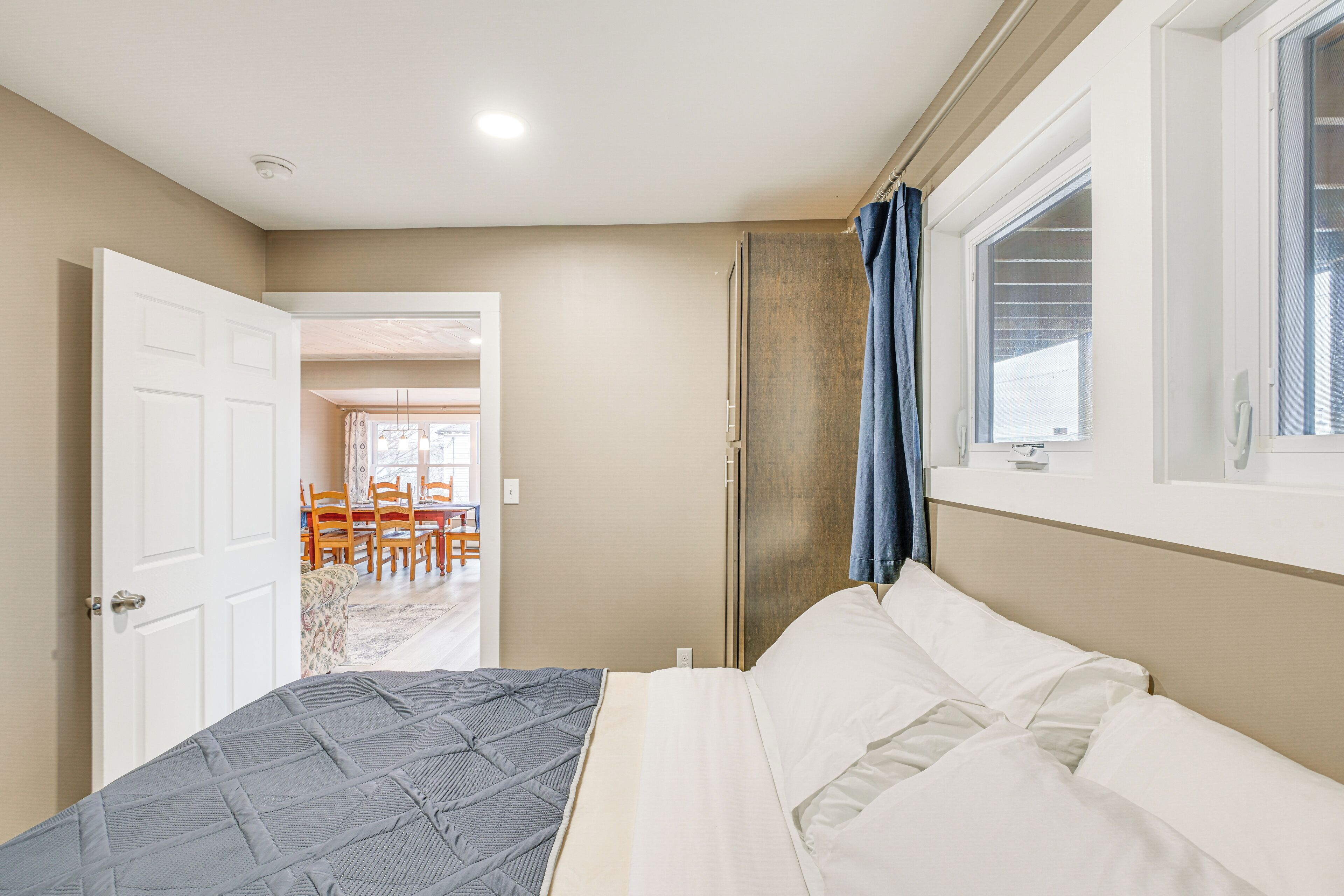 Una camera da letto con un letto, una finestra con tende, una porta che dà su una zona pranzo e una lampada a soffitto.