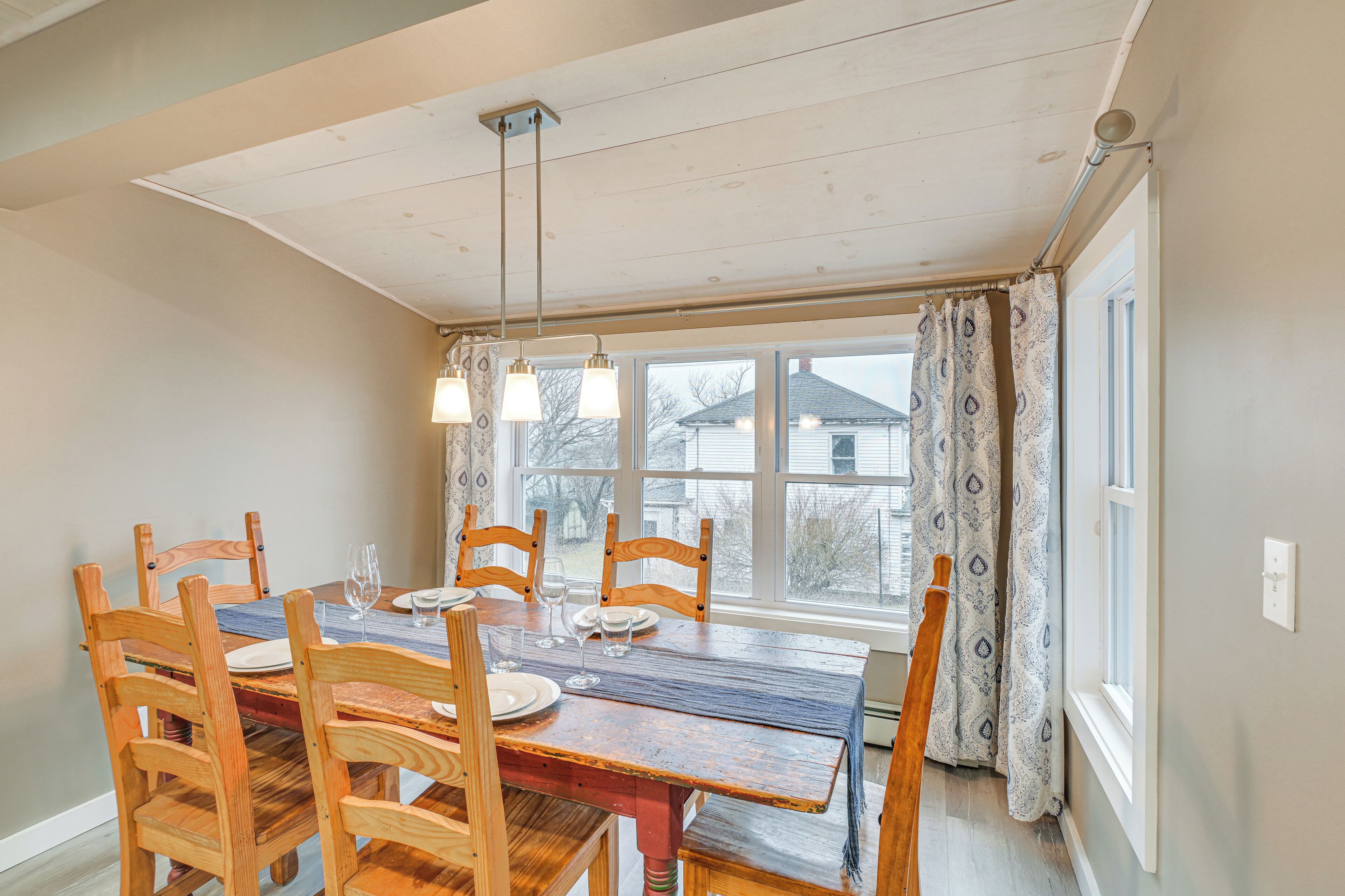 A dining room with a wooden table and chairs, a chandelier, and a view of houses through the window.