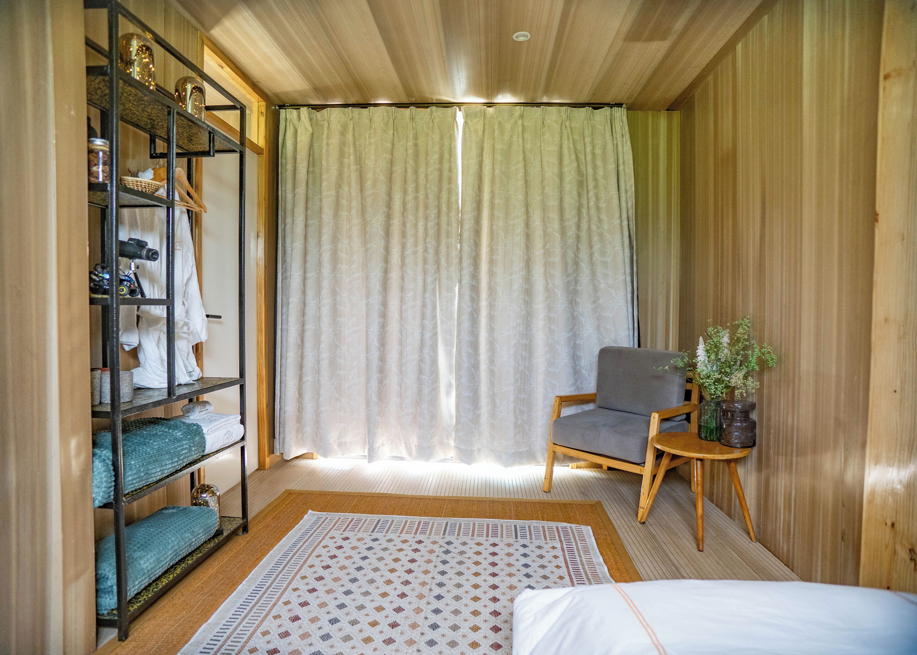 Una habitación con una cama, una silla, una estantería con ropa y una ventana con cortinas.