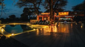 Un área junto a la piscina con maderas, sillones de descanso y un edificio con iluminación cálida.