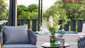 Un área de estar al aire libre con lámparas colgantes de mimbre, una silla con cojín gris y una mesa con un jarrón de flores.
