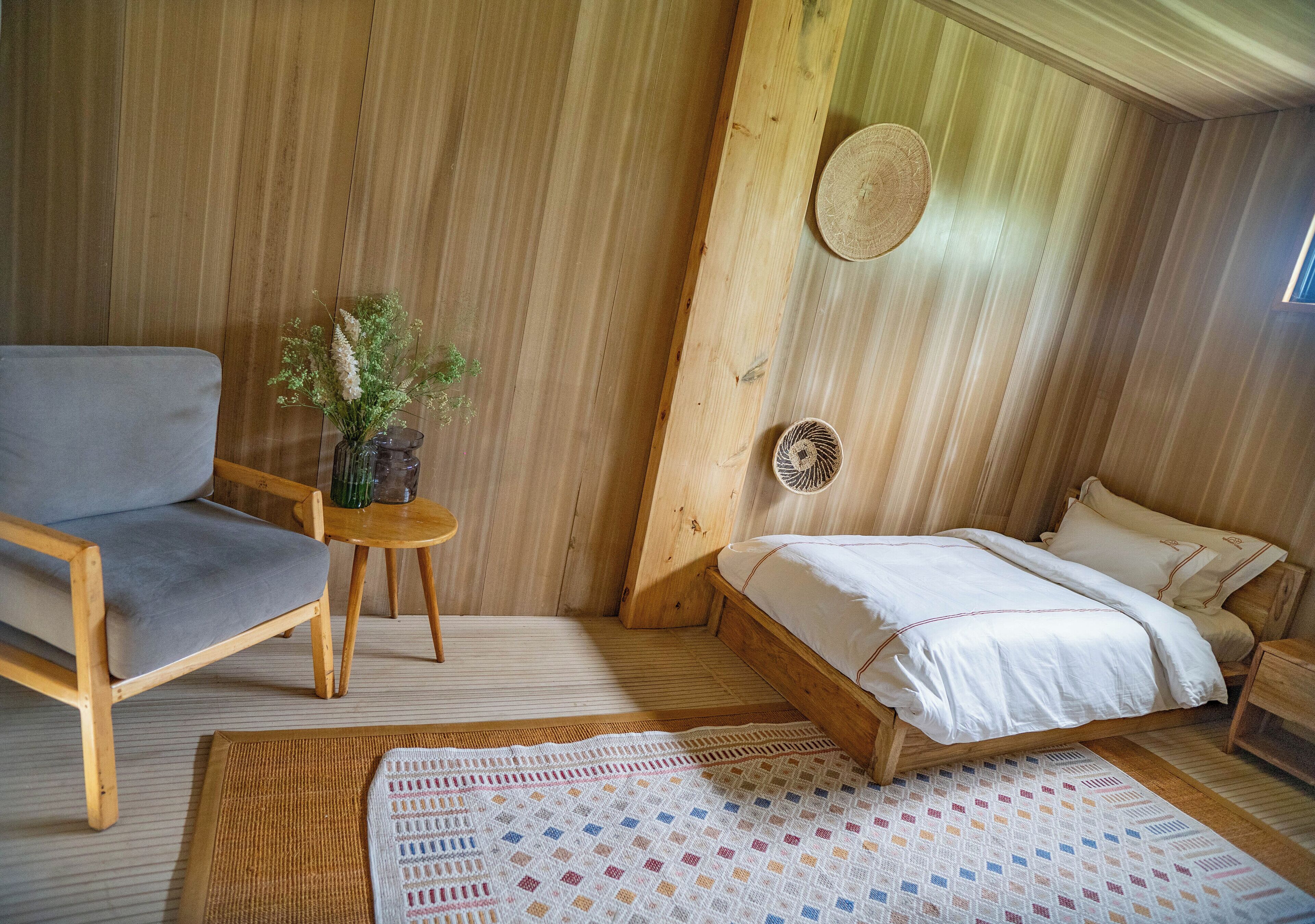 Una habitación de madera con una cama, una silla, una pequeña mesa con un florero y un tapete tejido en el suelo.