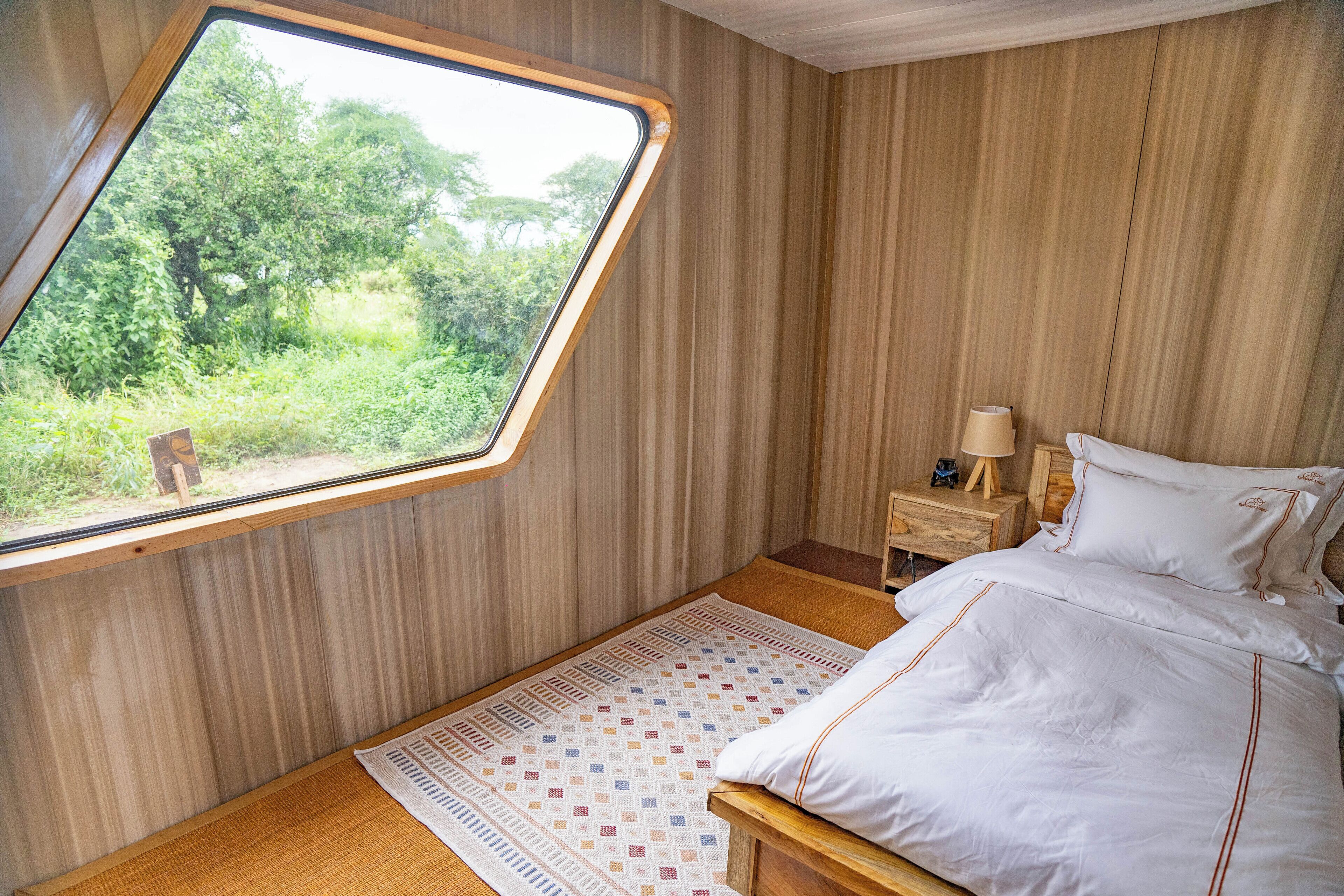 A bedroom with a wooden slatted wall, a window with a view of greenery, a bed with white bedding, and a small bedside table with a lamp.