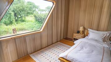 A bedroom with a wooden slatted wall, a window with a view of greenery, a bed with white bedding, and a small bedside table with a lamp.