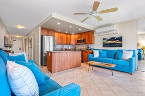 A modern living room with a blue sofa, a wooden coffee table, and a kitchen area with wooden cabinets and stainless steel appliances.
