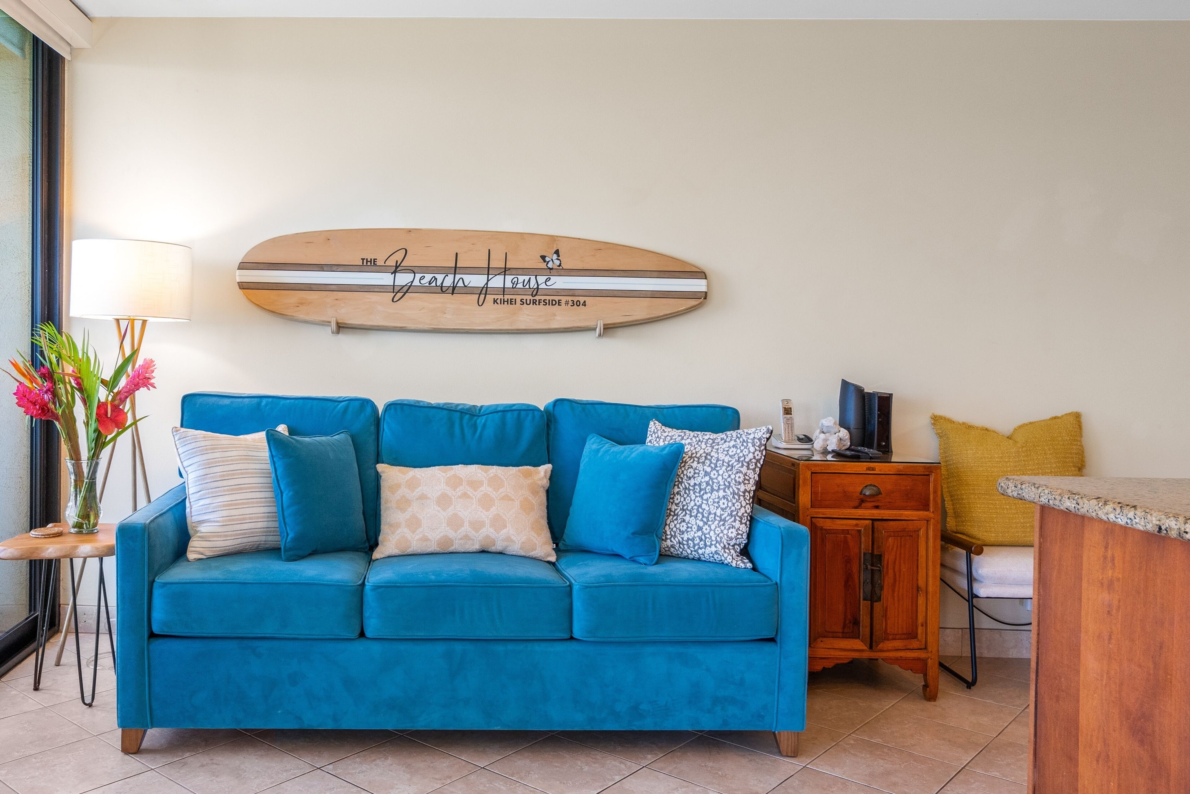 Un sofá azul con varios cojines, un adorno de pared de madera con texto, una lámpara y una mesita con un florero.
