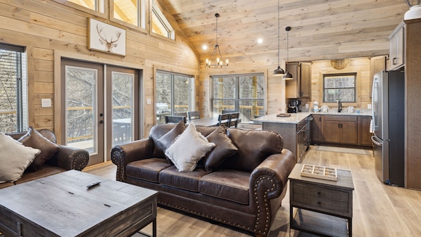 A spacious living room with a brown leather sofa, a wooden coffee table, and a dining area with a large window. - Tennessee Whiskey 3 Bedroom Cabin by RedAwning (Sevierville)