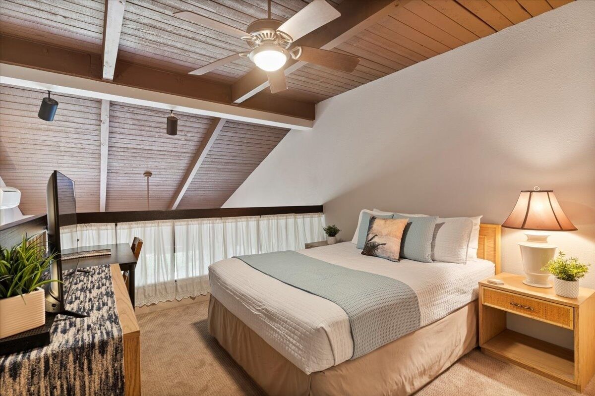Un dormitorio con techo inclinado, una cama con colcha en tonos claros, una mesita de noche con lámpara y un ventilador de techo.