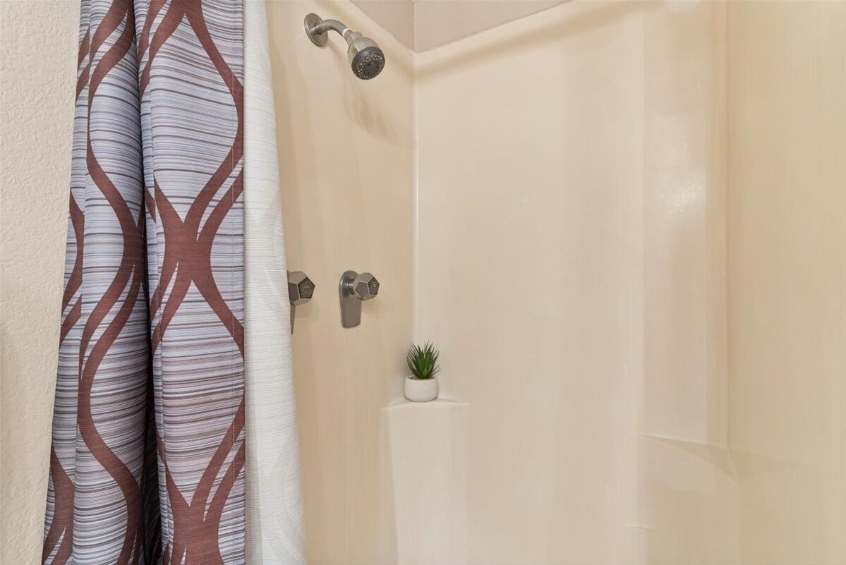 Ein Badezimmer mit beiger Wandfliese, einer Dusche mit Duschbrausehandstück und einer kleinen Topfpflanze an der Wand.