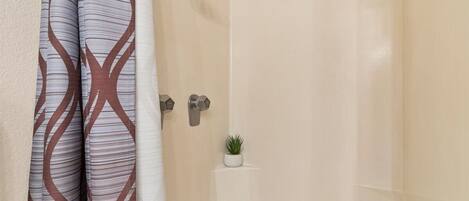 Ein Badezimmer mit beiger Wandfliese, einer Dusche mit Duschbrausehandstück und einer kleinen Topfpflanze an der Wand.