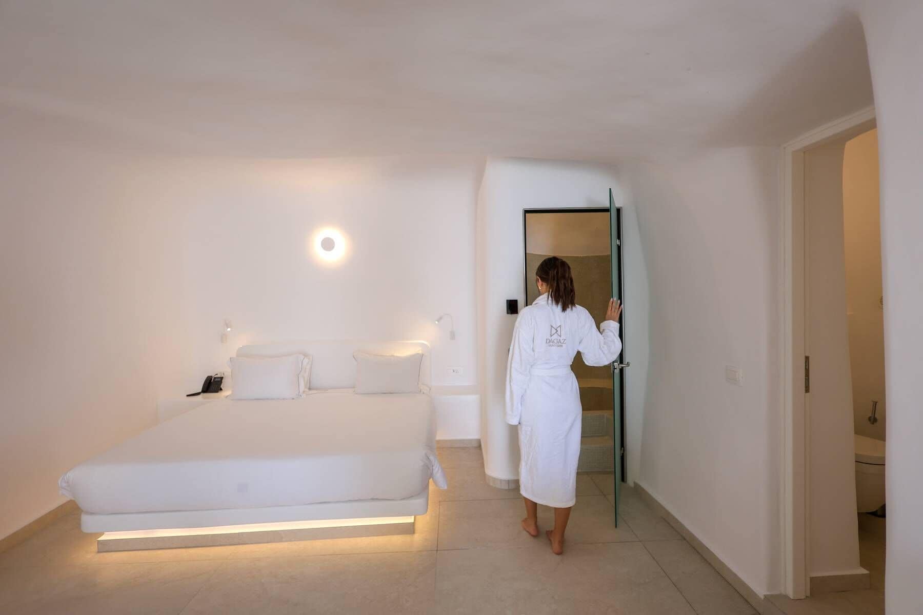 Une chambre à coucher comprenant un lit, une table de chevet, une lampe fixée au mur et une porte donnant sur une autre pièce.