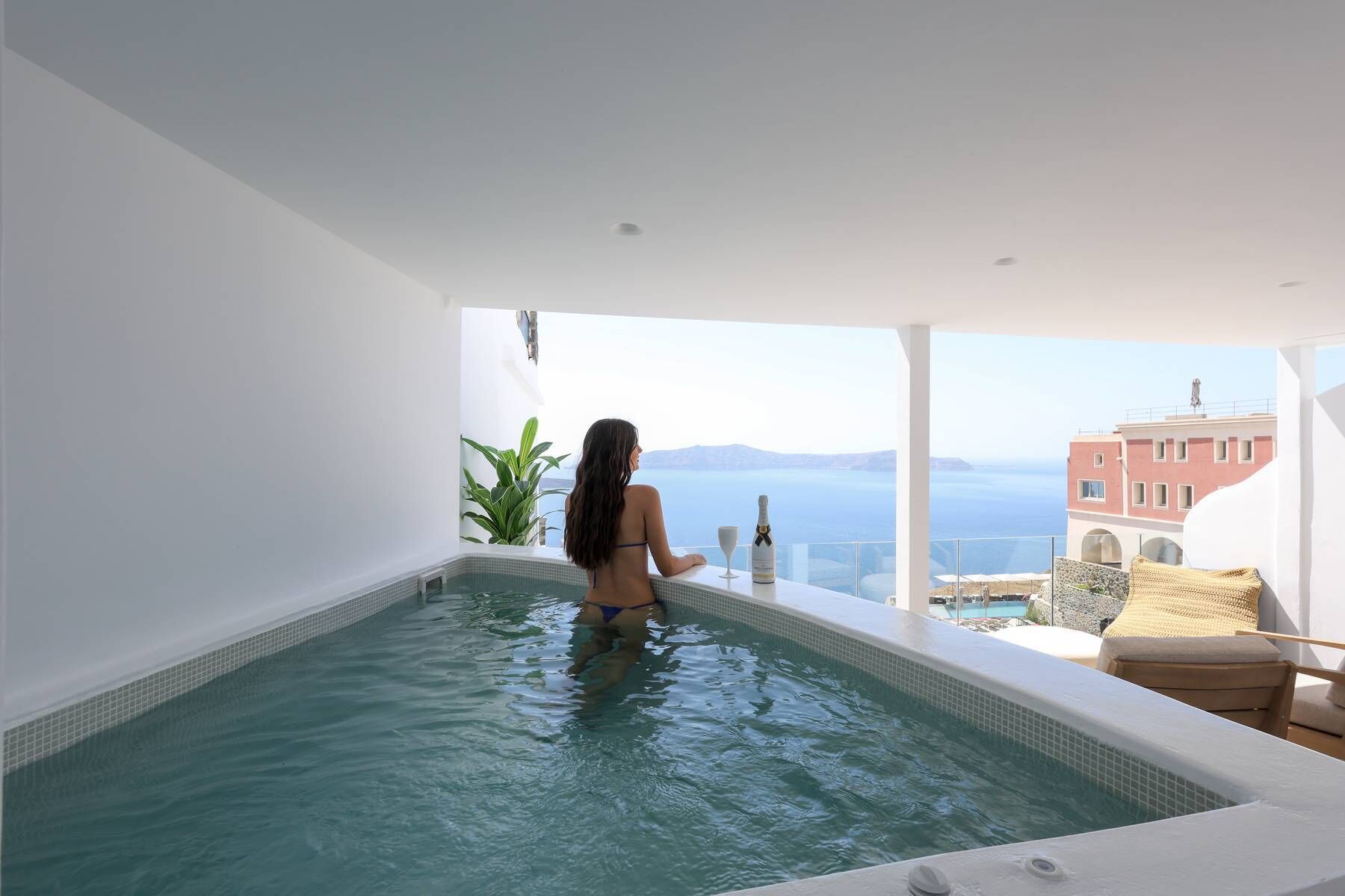Uma mulher em banheira, com uma garrafa de champanhe e um copo, apreciando uma piscina e a vista do mar e prédios.