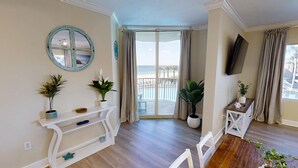A spacious room with a wooden floor, a white console table with potted plants, a round mirror, and a large sliding glass door leading to a balcony with a pool and lounge chairs. - Newly Renovated Beachfront w/ Coastal Style! Free Golf & Daily Activities! (Panama City Beach)